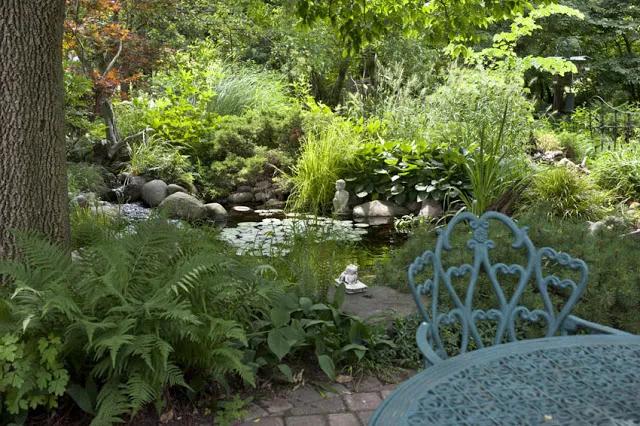A wrought iron table and chair lends an elegant touch to a brick patio lined with frothy ferns. Sunlight dapples on the pond's surface just beyond.