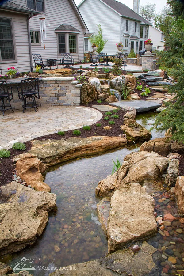 Stepping stones descend from the patio in the background, winding their way to the lower level, inviting visitors to explore the twists and turns of the stream and waterfalls.