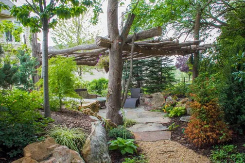 A rustic tree-type pergola provides shade on hot summer days in this beautiful backyard.